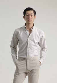 Camisa blanca de botones con rayas verticales en gris claro, corte ajustado, cuello puntiagudo y mangas largas. Se lleva con pantalones beige.