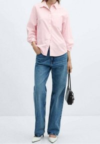 Helles rosa Hemd mit langen Ärmeln, kombiniert mit blauen, weiten Jeans. Das Model hält eine schwarze Handtasche und trägt weiße Absätze.