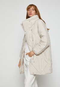 Lys beige pufferjakke med høj krave, diamant-quiltingmønster og trykknapper. Har lange ærmer og en afslappet pasform.