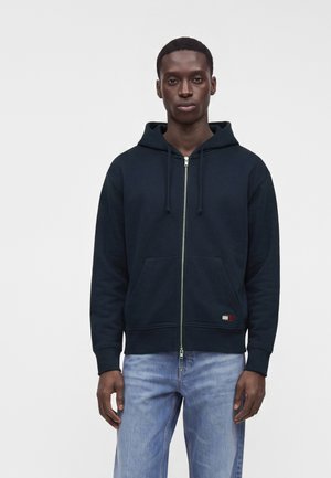 Giovane uomo che indossa una felpa nera con zip e jeans azzurri chiari, in piedi davanti a uno sfondo bianco semplice.