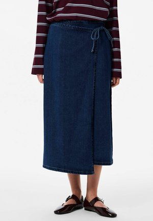 Ženska nosi temno modro prevezo iz jeansa, privezano okoli pasu, z bordočasto črtasto majico z dolgimi rokavi in temno rjavimi čevlji Mary Jane.