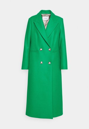 Kvinde iført en lang grøn dobbeltradet frakke, leopardmønstret bluse, tanfarvet lædernederdel, sorte støvler til knæet, holder en matchende grøn håndtaske.