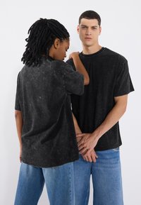 Două persoane poartă tricouri negre supradimensionate cu un efect de spălare, asortate cu blugi largi albaștri. Materialul prezintă un finisaj texturat.