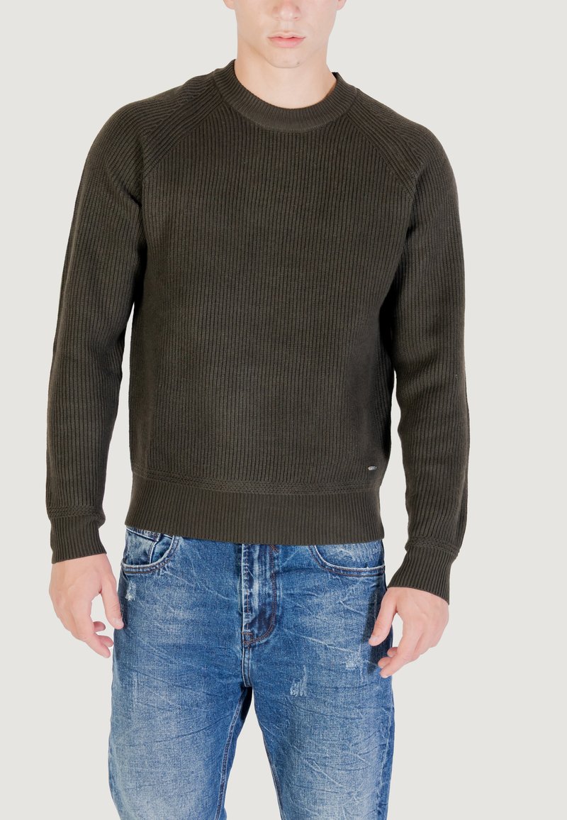 Uomo che indossa un maglione a coste verde scuro e jeans blu sbiaditi, in piedi contro uno sfondo chiaro e semplice, con le mani rilassate lungo i fianchi.