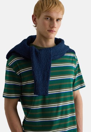 Junger Mann mit kurzem blondem Haar, der ein grünes, weißes und blau gestreiftes T-Shirt trägt, über dessen Schultern ein marineblauer Pullover gelegt ist.