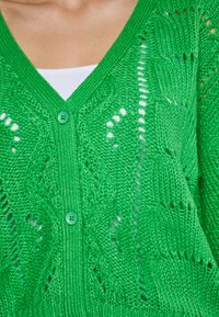 Grüner Strickcardigan mit V-Ausschnitt, zwei grünen Knöpfen, Lochmustern und strukturierten Details im gesamten Design. Leichter Stoff.