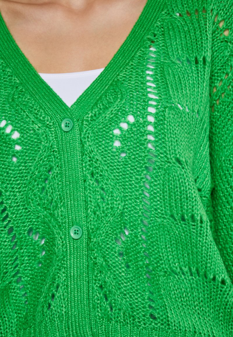 Grüner Strickcardigan mit V-Ausschnitt, zwei grünen Knöpfen, Lochmustern und strukturierten Details im gesamten Design. Leichter Stoff.