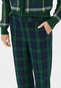 Pantaloni a quadri in verde scuro e blu navy, caratterizzati da una texture liscia e tasche laterali. Il motivo include righe alternate di dimensioni variabili.