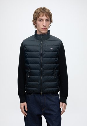 Jeune homme aux cheveux bouclés porte un gilet matelassé noir par-dessus une chemise noire à manches longues et un jean bleu, debout devant un fond blanc uni.