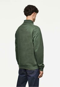 Zielony polar z wysokim kołnierzem, ściągaczami na rękawach i u dołu, oraz prostokątnym detalem akcentującym z tyłu. Połączony z niebieskimi dżinsami.