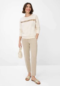 Creme sweatshirt med en glitrende vandret stribe, beige skræddersyede bukser og gyldne slidssandaler. Holder en beige håndtaske.