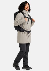Giacca leggera di colore crema con zip frontale, cappuccio regolabile e zaino nero. Abbinata a pantaloni scuri e scarpe nere strutturate.