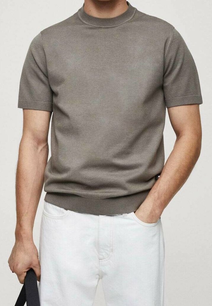 Homme portant un haut en maille gris à manches courtes et un pantalon blanc, une main dans la poche et l'autre tenant une sangle noire.