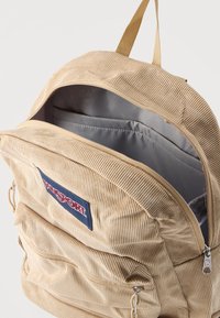 Μπεζ σακίδιο πλάτης από κορντόρι με μπροστινή τσέπη με φερμουάρ, πατζάμι με το λογότυπο Jansport, ανοιχτό κεντρικό διαμέρισμα και μπεζ ύφασμα για χερούλι.