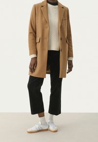 Manteau en laine marron avec deux boutons, manches longues et poches avant, superposé sur un pull en tricot torsad�é crème et un pantalon en velours côtelé noir, associé à des baskets blanches.