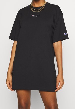 Osoba nosząca oversize czarną sukienkę w stylu t-shirt Champion z logo na piersi i rękawie, dopełnioną warstwowymi złotymi łańcuszkami.