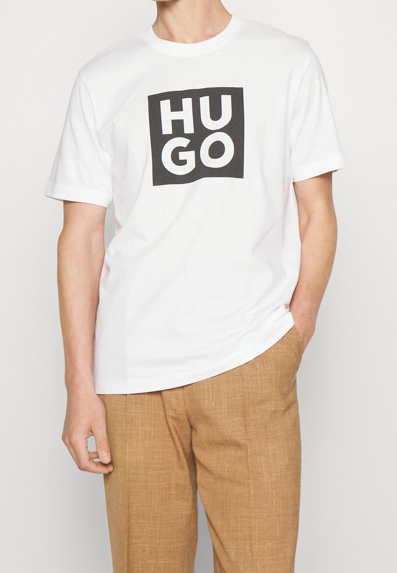 T-shirt blanc à manches courtes avec un logo carré noir audacieux affichant le mot "HUGO" en blanc. Le tissu semble lisse et léger.