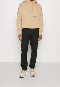 Sweatshirt beige à capuche avec poche avant et logo blanc, associé à un pantalon noir et des chaussures de sport beiges. Matériau lisse, coupe décontractée.