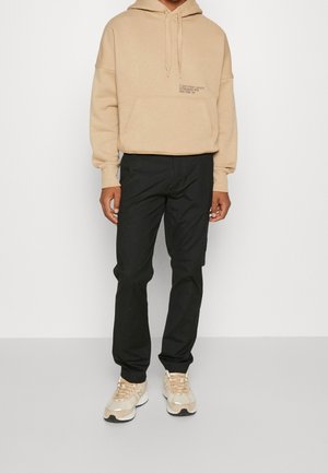 Beige Kapuzenpullover mit Fronttasche und weißem Logo, kombiniert mit schwarzen Hosen und beigefarbenen Sportschuhen. Glattes Material, lockere Passform.