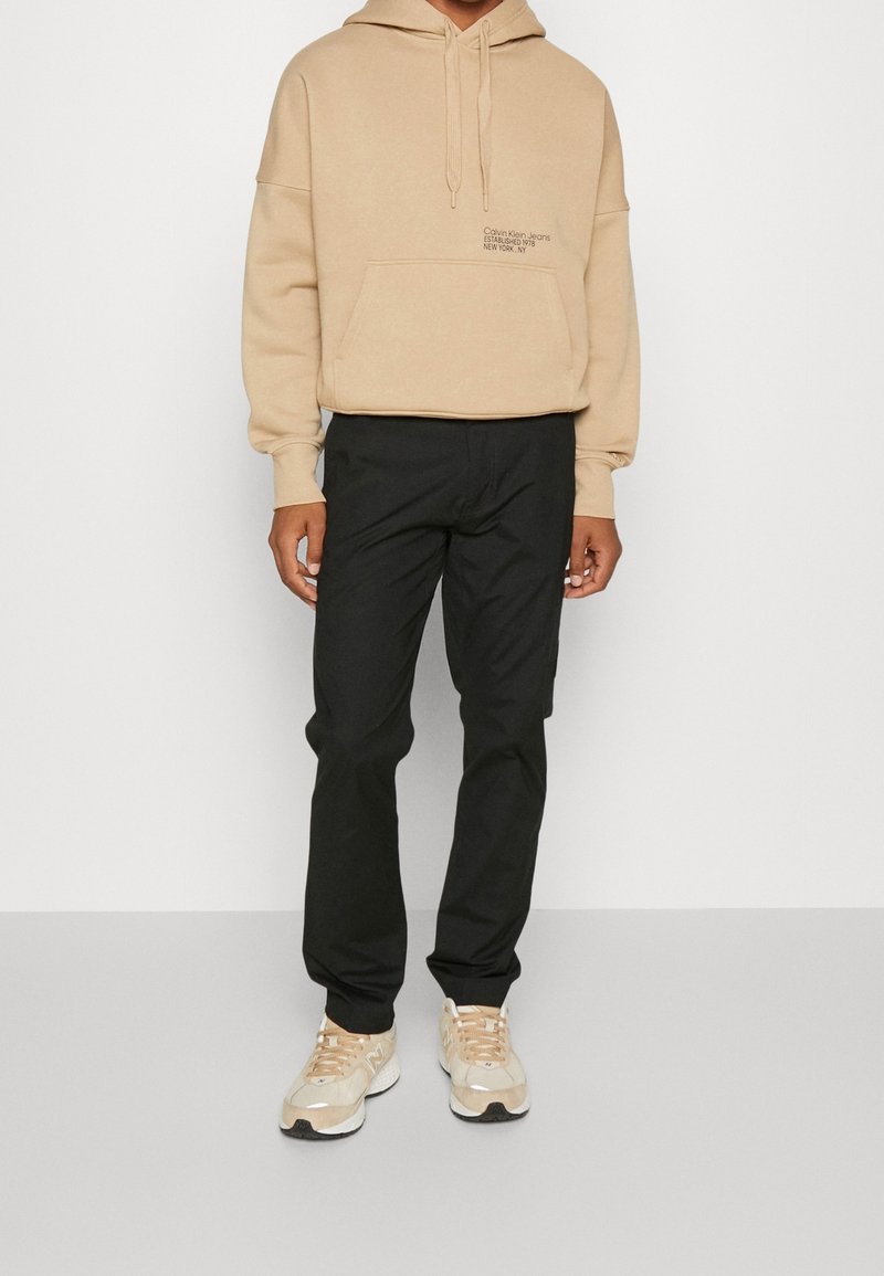 Sweatshirt beige à capuche avec poche avant et logo blanc, associé à un pantalon noir et des chaussures de sport beiges. Matériau lisse, coupe décontractée.
