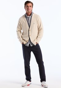 Jonge man die met zijn handen in zijn zakken staat, gekleed in een crèmekleurige kabelgebreide cardigan, gestreepte shirt, donkere broek en witte sneakers met rode accenten.