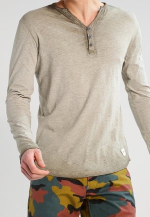 Camisa henley de manga larga en verde oliva claro con tres botones y escote redondeado. Se lleva con pantalones de camuflaje multicolor.