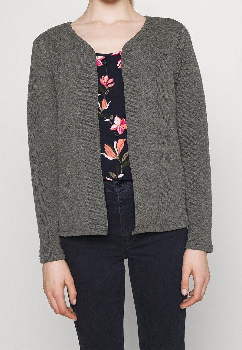 Femme portant un cardigan gris texturé ouvert sur le devant, par-dessus un haut bleu marine à imprimé floral rose et un pantalon bleu foncé.