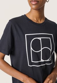 Juodas medvilnės marškinėlis su baltu geometriniu logotipu. Apykaklė su apvaliu iškirpimu ir trumpomis rankovėmis. Lygus tekstūros paviršius su laisvu siluetu.