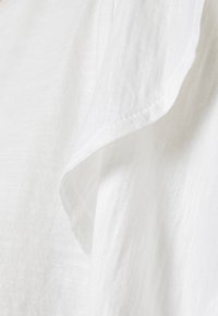 Vit, lättviktig tyg med en texturerad yta och en något draperad halsringning. Kanten har en prydlig sömslutning.