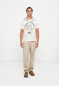 Junger Mann steht, trägt ein weißes T-Shirt mit Grafik, beige Hose, braune Schuhe, mit einem gestreiften Pullover über den Schultern vor einem schlichten weißen Hintergrund.