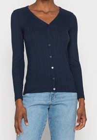 Kvinna iklädd en åtsittande marinblå ribbad knappad kofta med långa ärmar och V-ringning, kombinerad med ljusblå jeans.