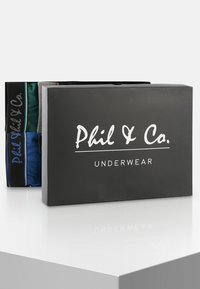 Černá krabice s nápisem "Phil & Co. SPODNÍ PRÁDLO" obsahuje čtyři páry vícebarevného spodního prádla: zelené, šedé, modré a černé s logovanými pásky.