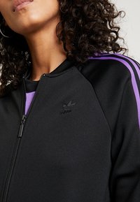 Svart zip-up jacka med lila detaljer, som har en ståkrage och broderad logotyp. Tillverkad av ett slätt, elastiskt material med en figurnära design.