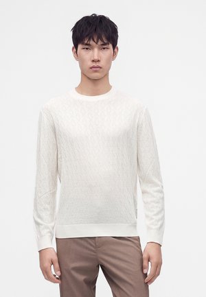 Asian man wearing a white textured long-sleeve sweater and brown pants standing against a plain light background.