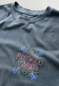 Blau-graues T-Shirt mit Rundhalsausschnitt, auf dem in einem pinkfarbenen Kreis der Schriftzug "Fleetwood Mac" steht, umgeben von blauen Blumen und grünen Blättern.