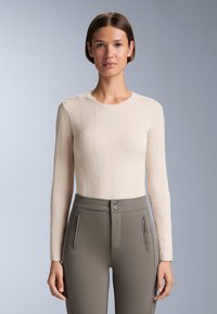 Beiges Langarmshirt mit Rautenmuster und rundem Ausschnitt, kombiniert mit hoch taillierter grauer Hose mit vorderen Reißverschlüssen und Zwei-Knopf-Verschluss.