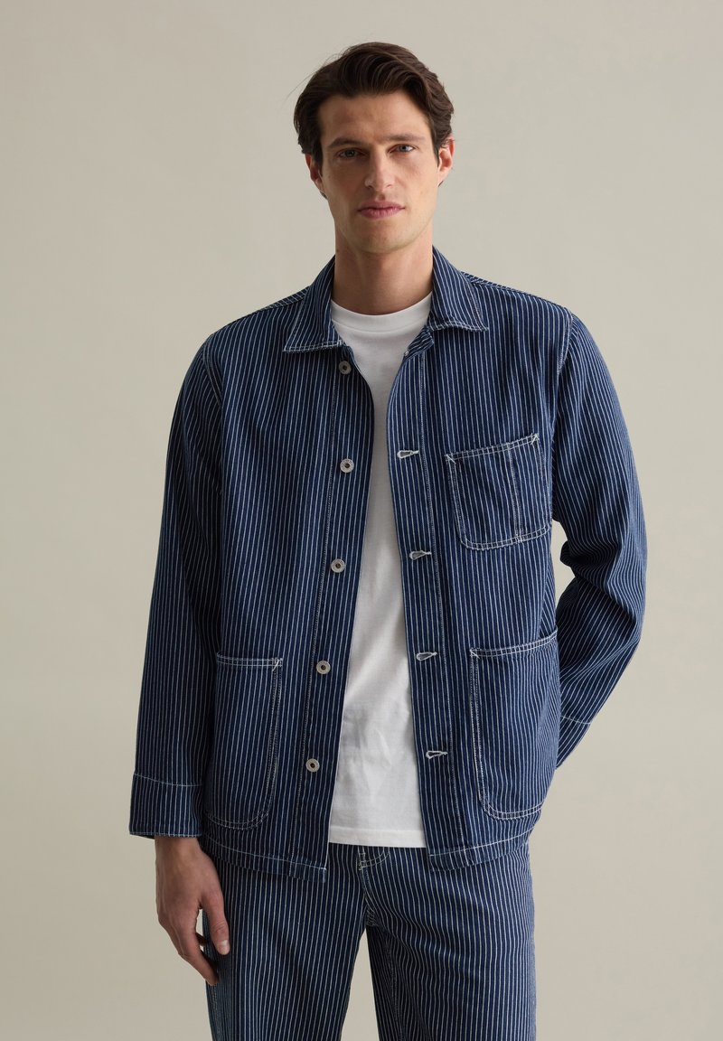 Blue striped work jacket made of cotton, featuring a button front, two front pockets, and contrasting white stitching. Worn over a white shirt.