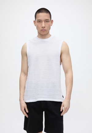 Jeune homme aux cheveux courts portant un haut blanc texturé sans manches et un short noir, debout devant un fond clair uni.