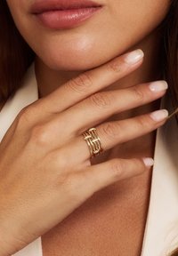 Goldfarbener Ring mit verflochtenen Bändern, von denen drei mit klaren Steinen akzentuiert sind. Glatte, polierte Oberfläche auf einer Hand.