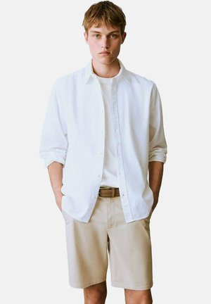 Witte overhemd met opgerolde mouwen, gedragen over een witte t-shirt, gecombineerd met beige shorts en een leren bruine riem. Eenvoudig en klassiek ontwerp.