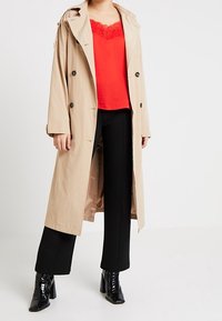 Trench coat beige con cappuccio, chiusura a bottoni e maniche lunghe. Indossato sopra una maglia rossa con dettagli in pizzo e pantaloni neri, abbinati a stivali con tacco lucidi.