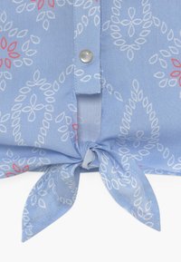 Tela azul con patrones florales en blanco y rojo, que presenta un botón de presión en la parte delantera y un nudo atado en el centro inferior.