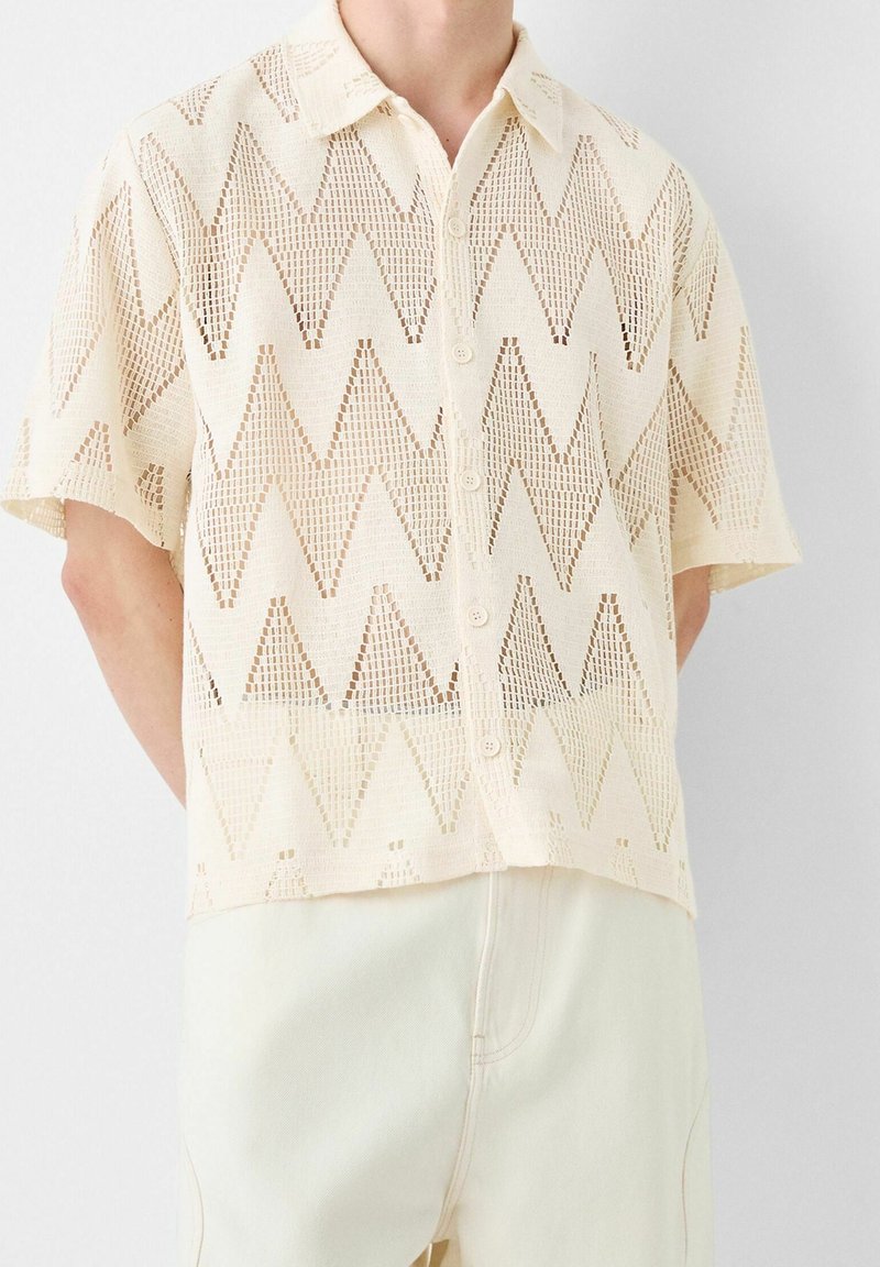 Maglietta a maniche corte color crema, lavorata a maglia, con un motivo a zigzag, chiusura con bottoni e vestibilità rilassata. Design aperto e testurizzato.