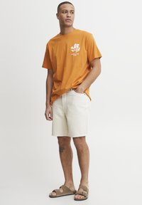 Camiseta de algodón naranja con gráfico floral blanco, combinada con pantalones cortos de mezclilla beige y sandalias marrones. Diseño simple y corte relajado.