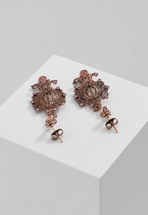 Para ozdobnych miedzianych kolczyków z misterną ażurową pracą, małymi przezroczystymi kamieniami i zapięciami na motylki, leżąca na białej powierzchni.