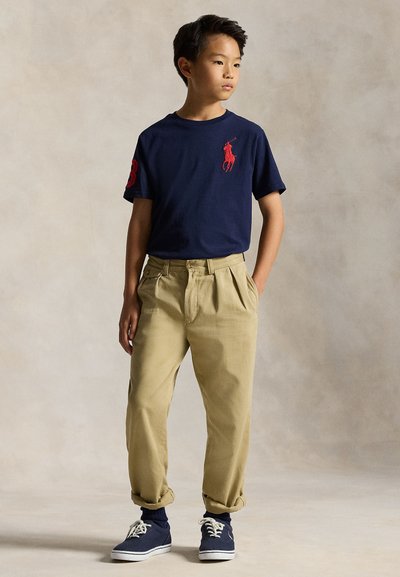 Poiss seisab käed taskutes, seljas tumesinine T-särk punase logoga, beežid üles kääritud püksid, tumesinised sokid ja tumesinised tossud.