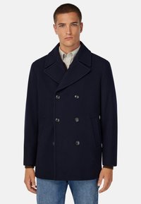 Marineblauer Wollpeacoat mit doppelt geknöpftem Verschluss, großen Revers, Seitentaschen und lockerer Passform, über einem leichten Hemd getragen.