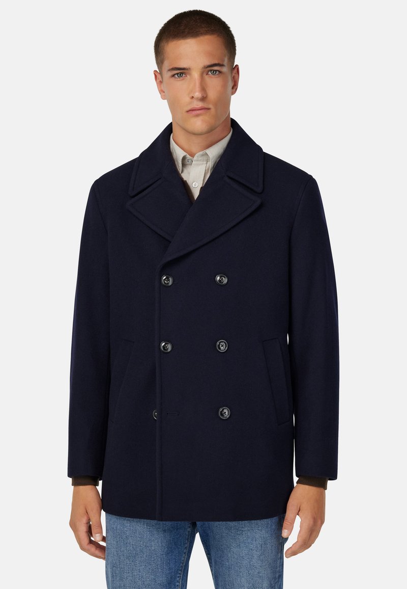 Marineblauer Wollpeacoat mit doppelt geknöpftem Verschluss, großen Revers, Seitentaschen und lockerer Passform, über einem leichten Hemd getragen.