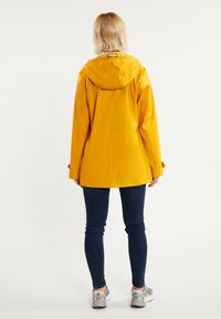 Chubasquero amarillo impermeable con capucha, que presenta un cierre frontal oculto y puños ajustables, combinado con unos vaqueros oscuros y zapatillas grises.