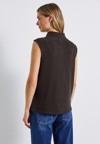 Ärmlös, mörkbrun texturerad topp med hög krage, med boxig passform och ribbat mönster, i kombination med blå jeans.